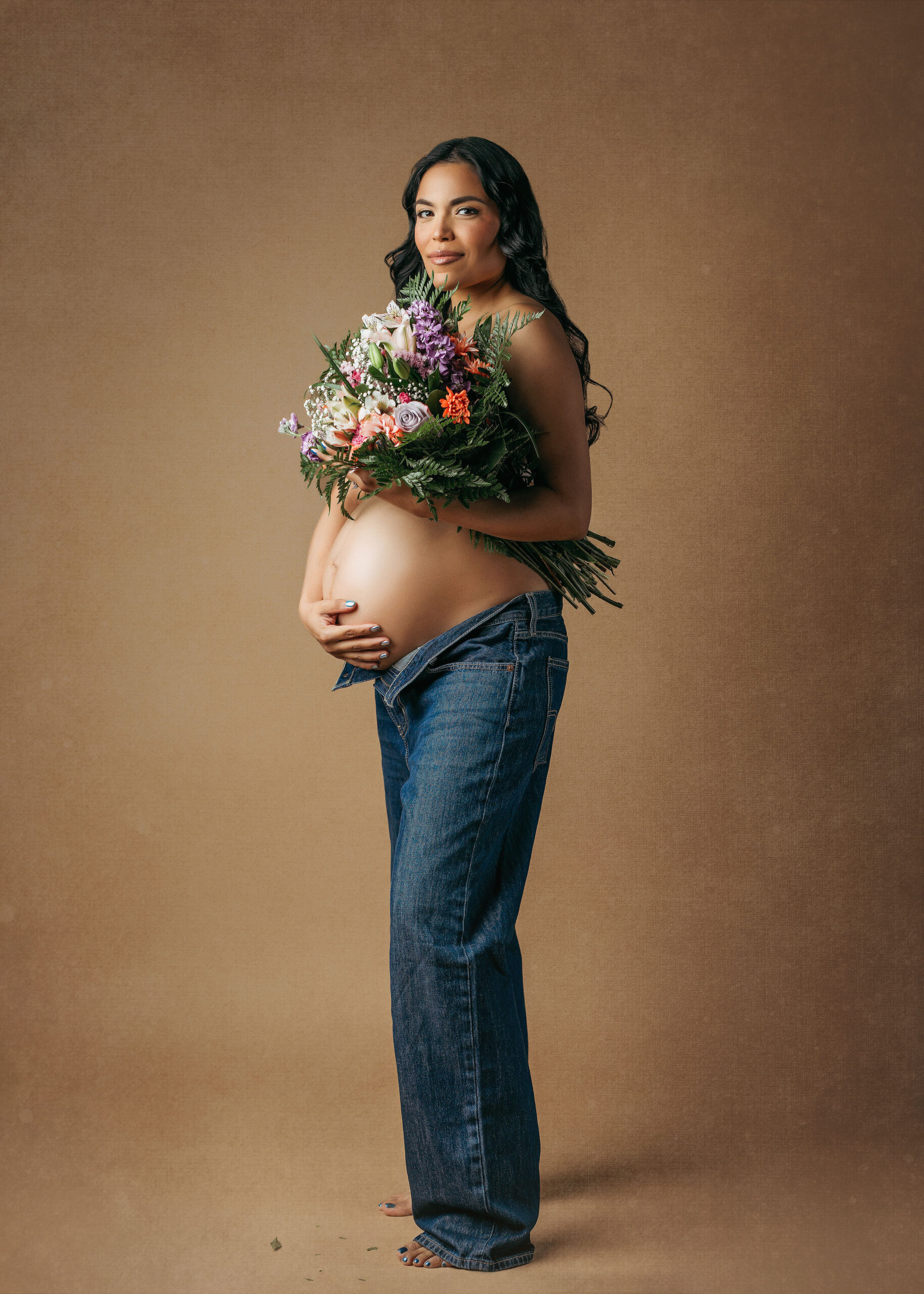 photo of pregnant woman standing wearing unzipped blue jeans, no top and holding a bouquet of flowers that hide her breasts