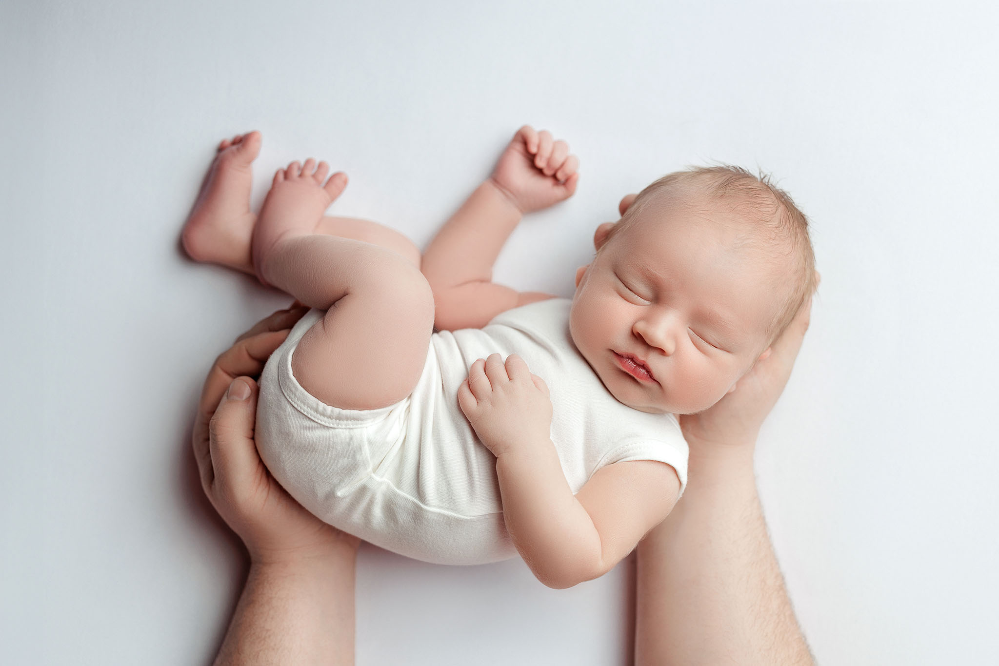 Baby boy wearing a white onsie posed in father's hands on white backdrop